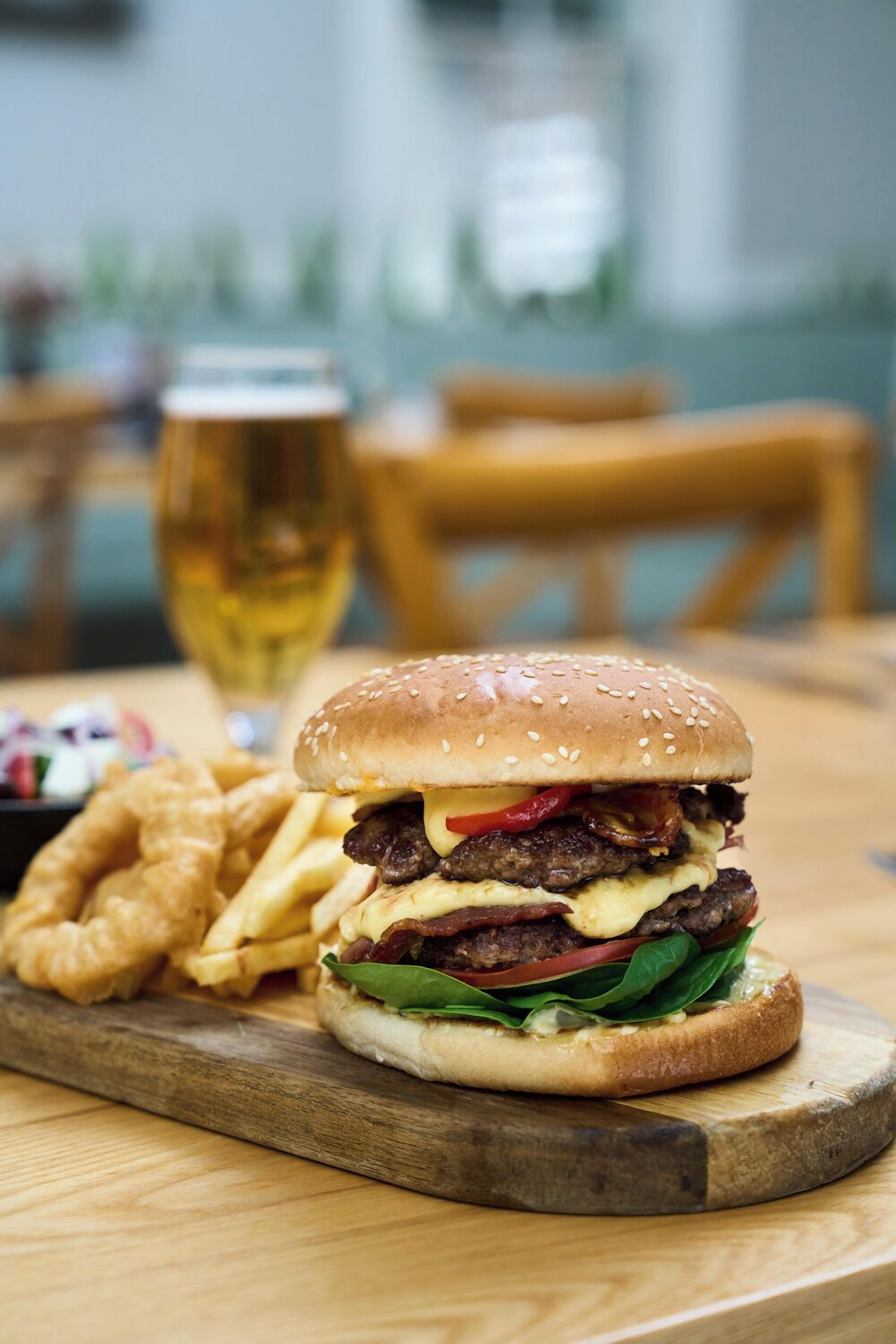 Una hamburguesa con papas fritas sobre una tabla de madera, una jarra de cerveza y una silla al fondo.