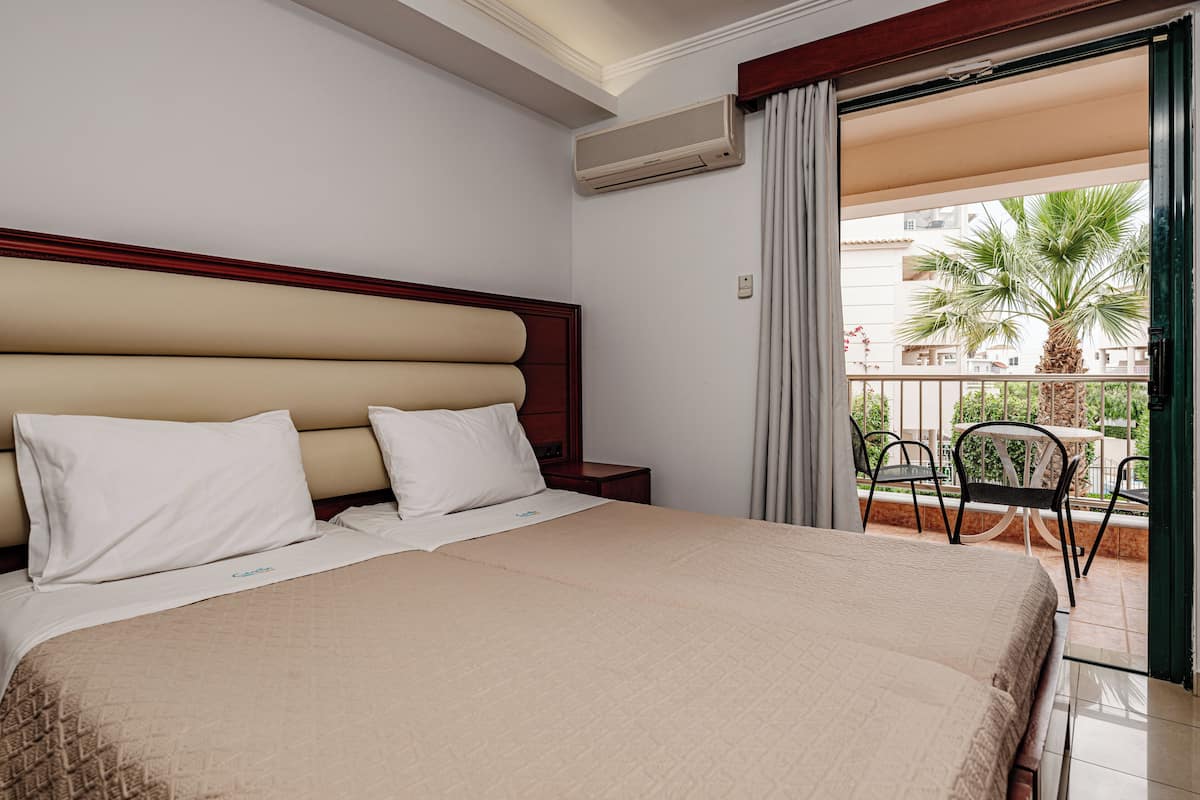 A hotel room with a large bed, a wooden headboard, a balcony with chairs, and an air conditioning unit.