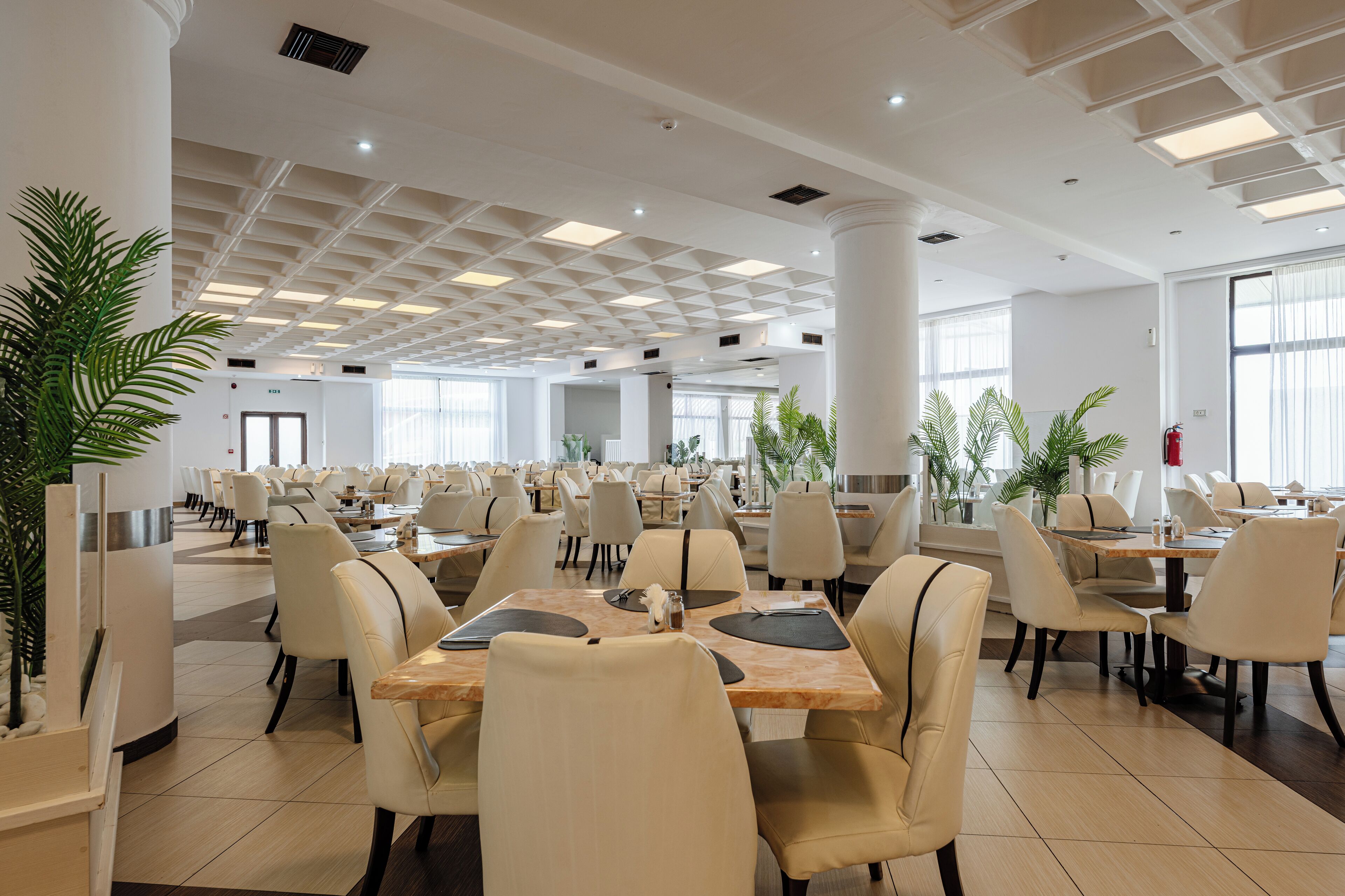 A spacious indoor dining area with multiple tables and chairs, large windows, and ceiling lights.