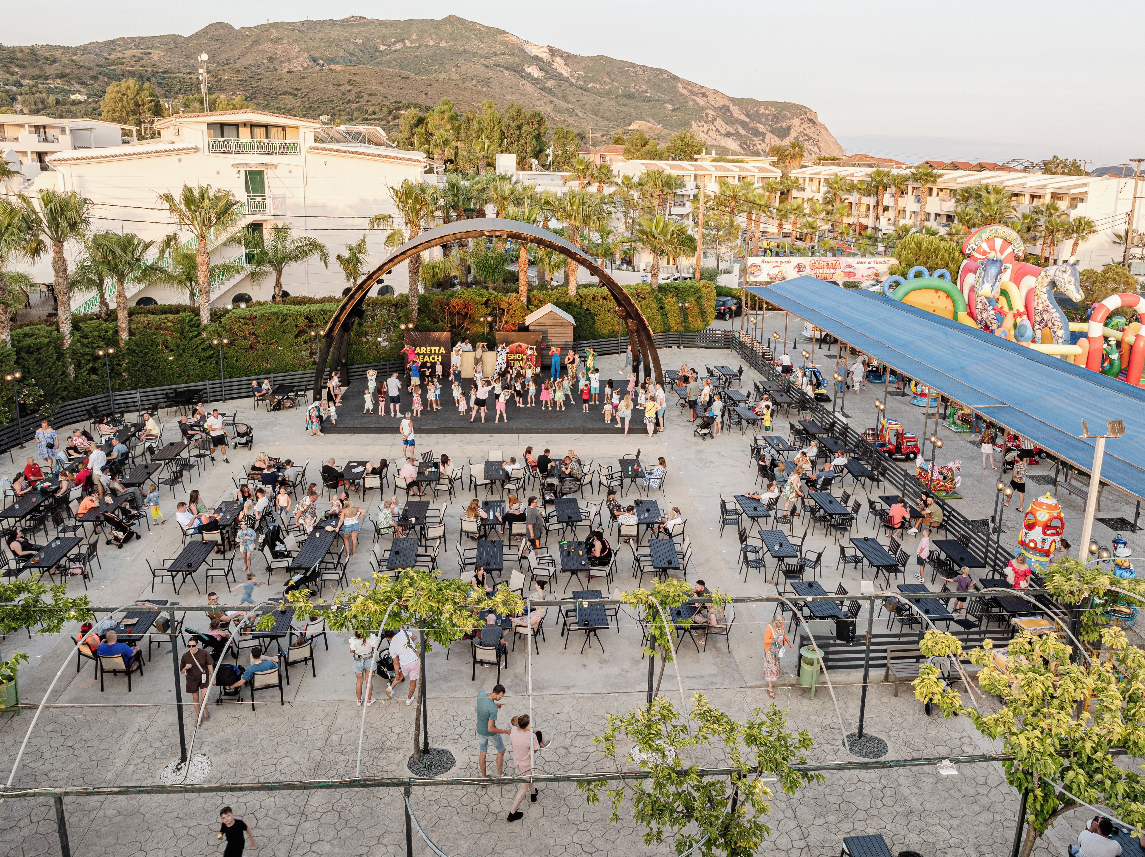 An outdoor seating area with tables and chairs, surrounded by palm trees and a stage for events.