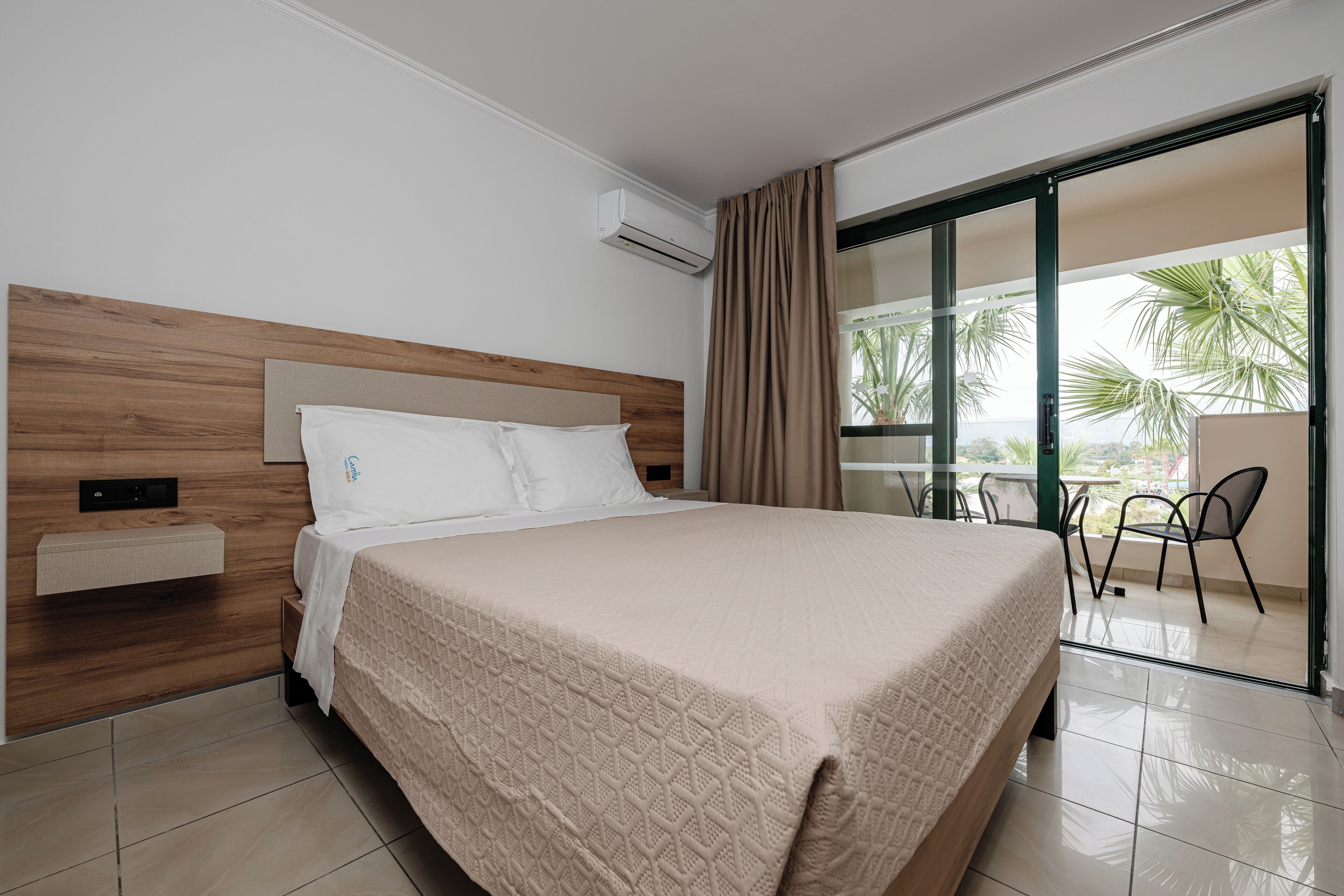 A hotel room with a large bed, a wooden headboard, a built-in shelf, and a sliding glass door leading to an outdoor area with chairs and a table.