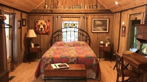 A cozy bedroom with a wooden ceiling, a bed with a colorful quilt, a nightstand with a lamp, a wooden bench, and a desk with a chair. - Holyfield Cabin and Bunkhouse (Flag Pond)
