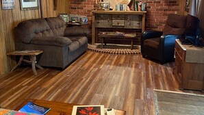 A cozy room with a wooden ceiling, a couch, a chair, a small table, and a bookshelf with various items. - Holyfield Cabin and Bunkhouse (Flag Pond)