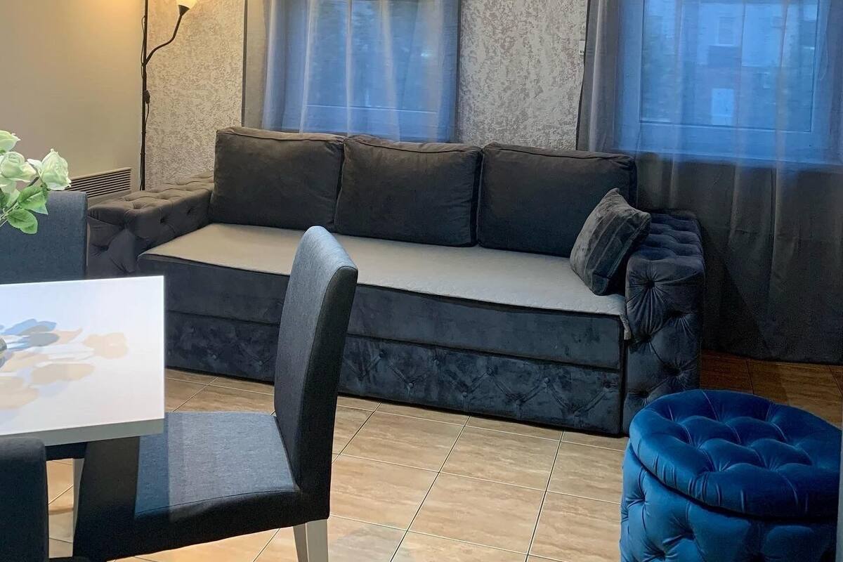 Una sala moderna con sofá, sillón, una otomana azul, lámpara y ventana con cortinas.