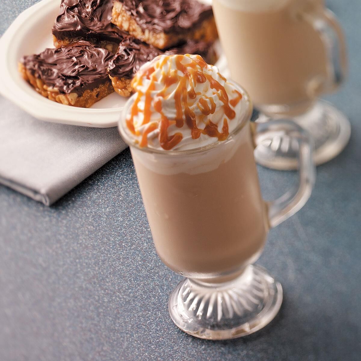 Bebida postre en taza de vidrio con crema batida y un hilo de caramelo, acompañada de pastelería cubierta de chocolate en un plato.
