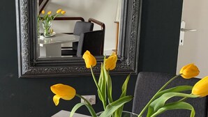 A white table with a vase of yellow tulips, a remote control, and a candle. A large mirror hangs above the table, reflecting a staircase and a chair. - Guesthouse Vladimir & Ana (Pancevo)