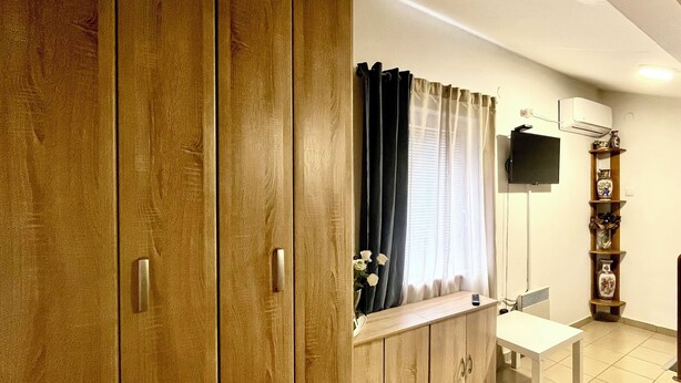 A room with wooden wardrobes, a white table, a TV on the wall, and a vase on a shelf. - Guesthouse Vladimir & Ana (Pancevo)