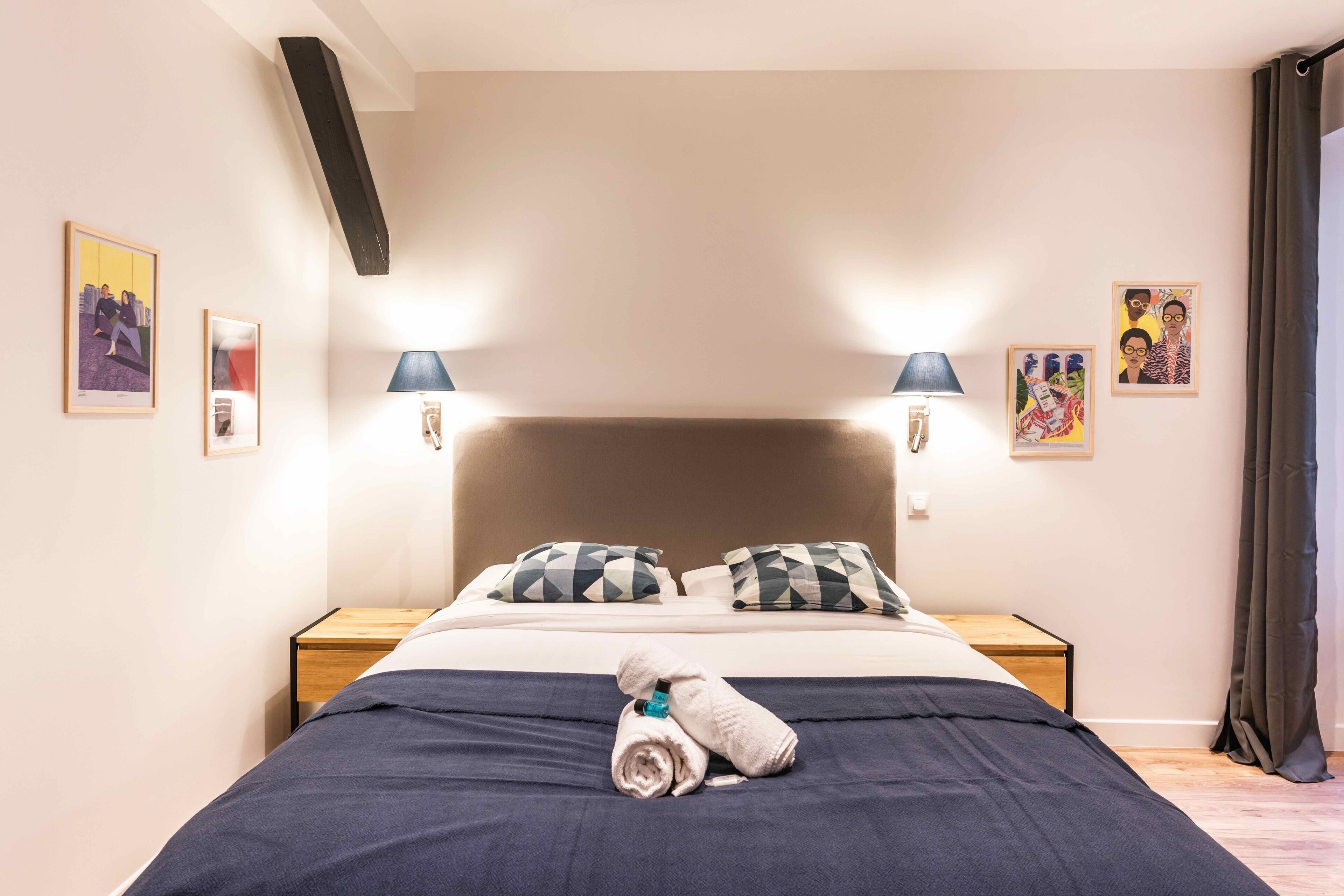 Una cama bien hecha con una colcha azul oscuro y dos almohadas, una mesita de noche de madera con una lámpara, y cuadros en las paredes.