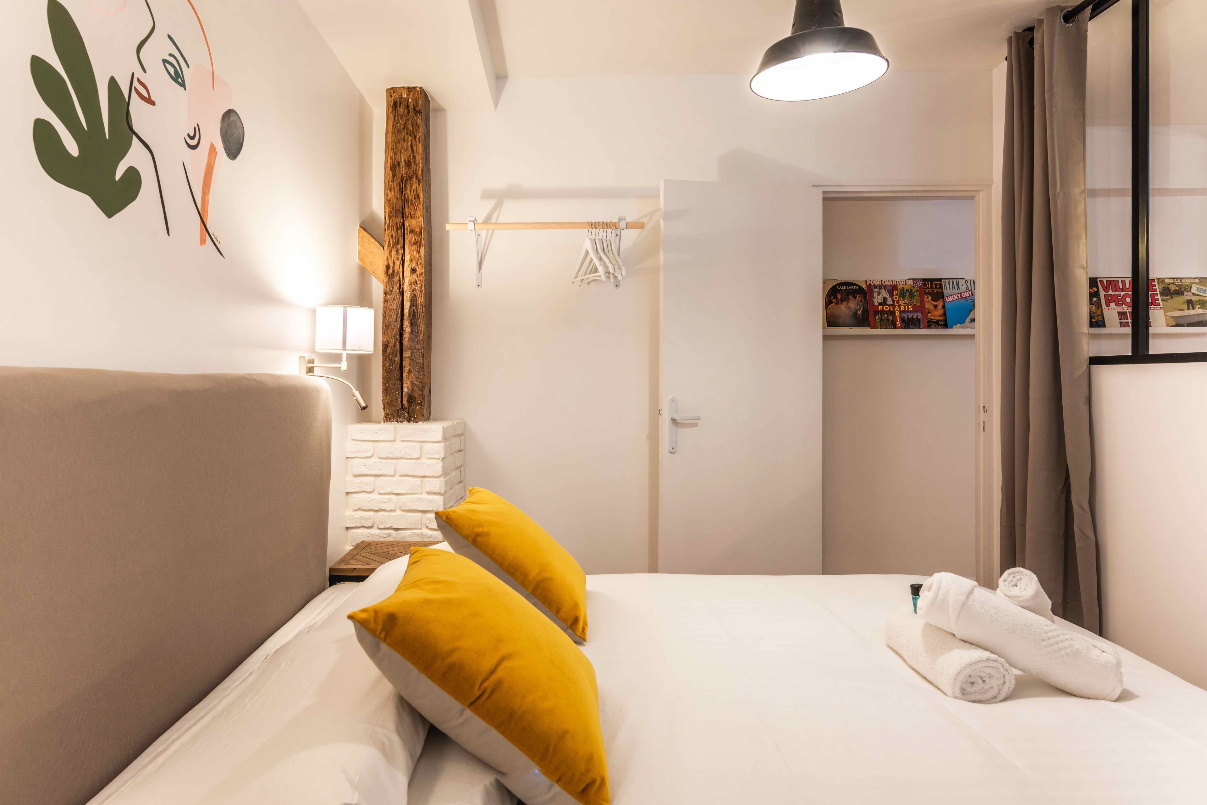 Camera d'albergo con un letto, due cuscini, un comodino, una lampada, una testiera in legno, un armadio e una porta che dà su un'altra stanza.
