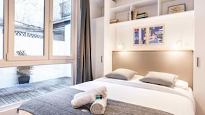 Une chambre d’hôtel avec un lit, une fenêtre, une plante et une étagère contenant des livres et une horloge.
