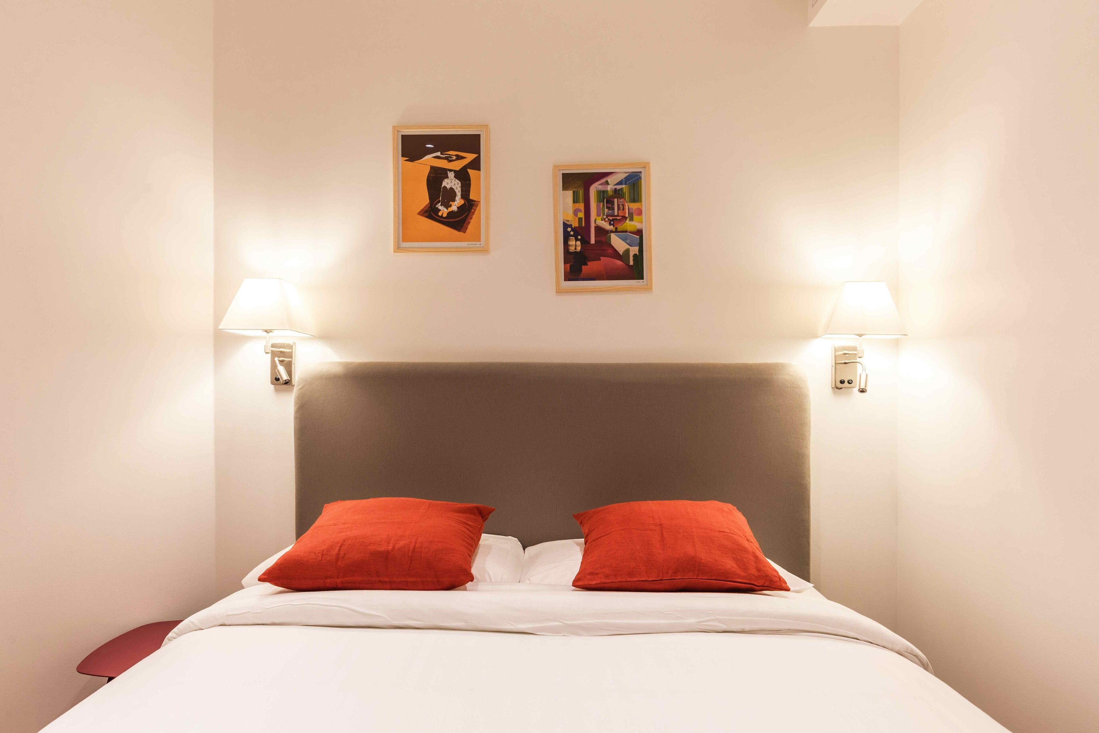 A hotel room with a large bed, two red pillows, a grey headboard, and two wall-mounted lamps.