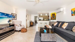 Ein modernes Wohnzimmer mit Sofa, Fernseher und Couchtisch. Im Hintergrund ist eine Küche mit Essbereich zu sehen.