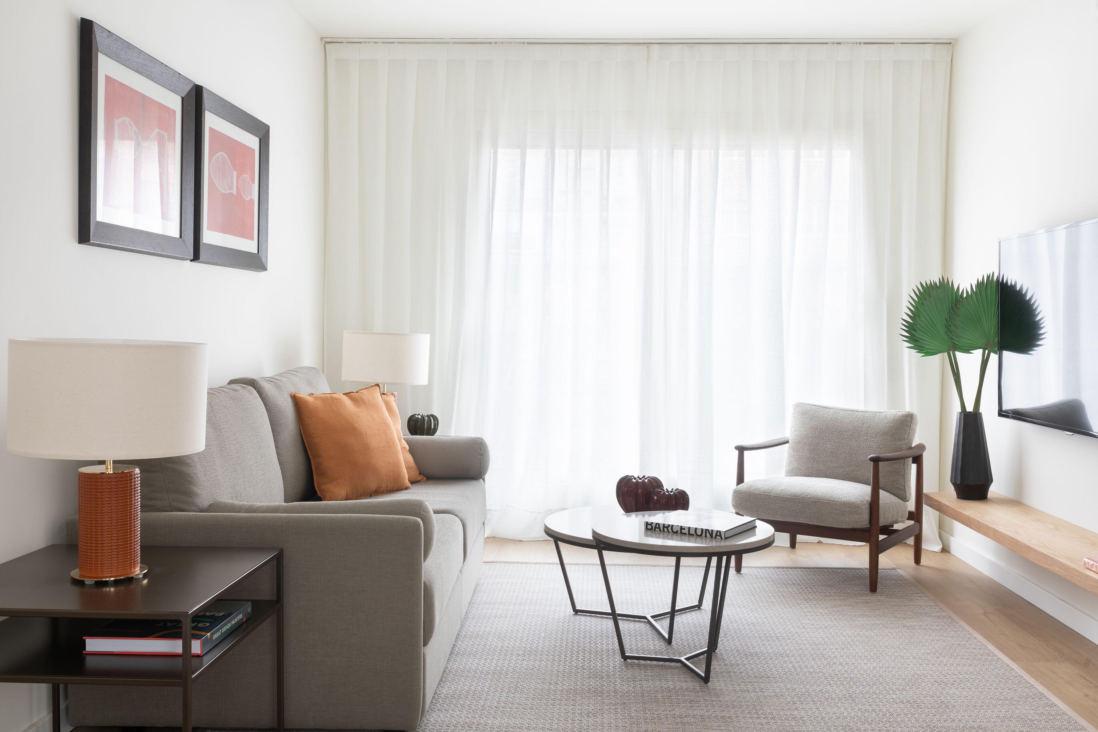 A modern living room with a grey sofa, a round coffee table, a bookshelf, a flat-screen TV, and a large window with sheer curtains.