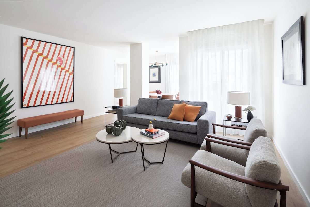 A modern living room with a grey sofa, two armchairs, a coffee table, and a bench. There is a large abstract painting on the wall and a lamp on a side table.