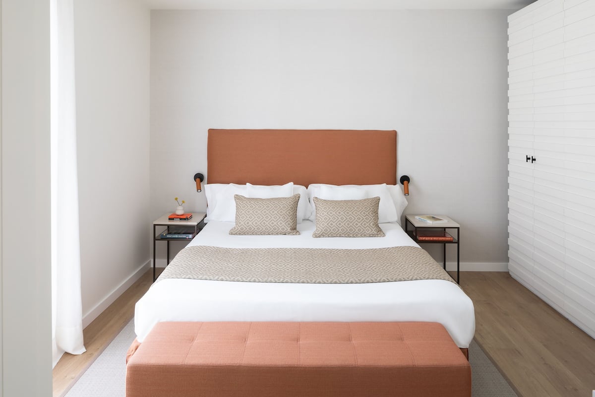 A bedroom with a bed, two bedside tables, a nightstand, a window with blinds, and a wooden floor.