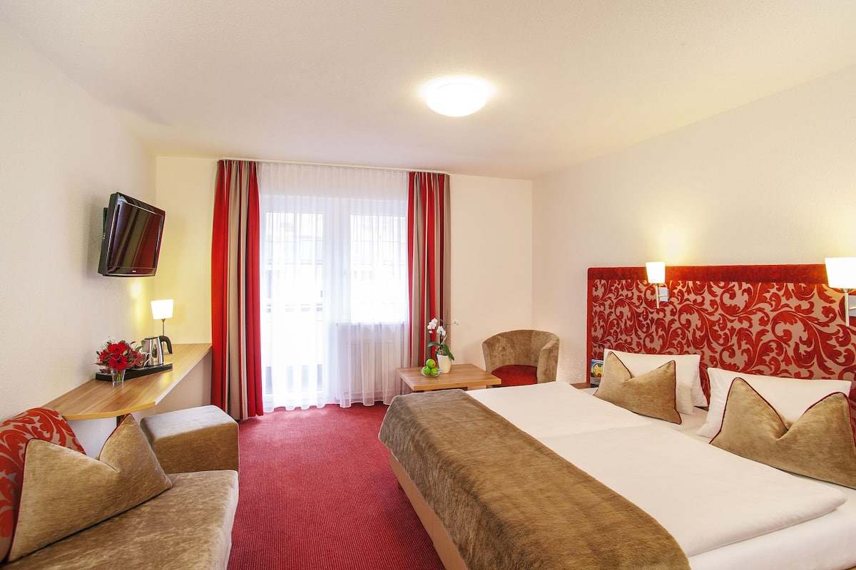 Una habitación de hotel con una cama, dos sillones, un escritorio con lámpara, un televisor y un ventanal con cortinas.