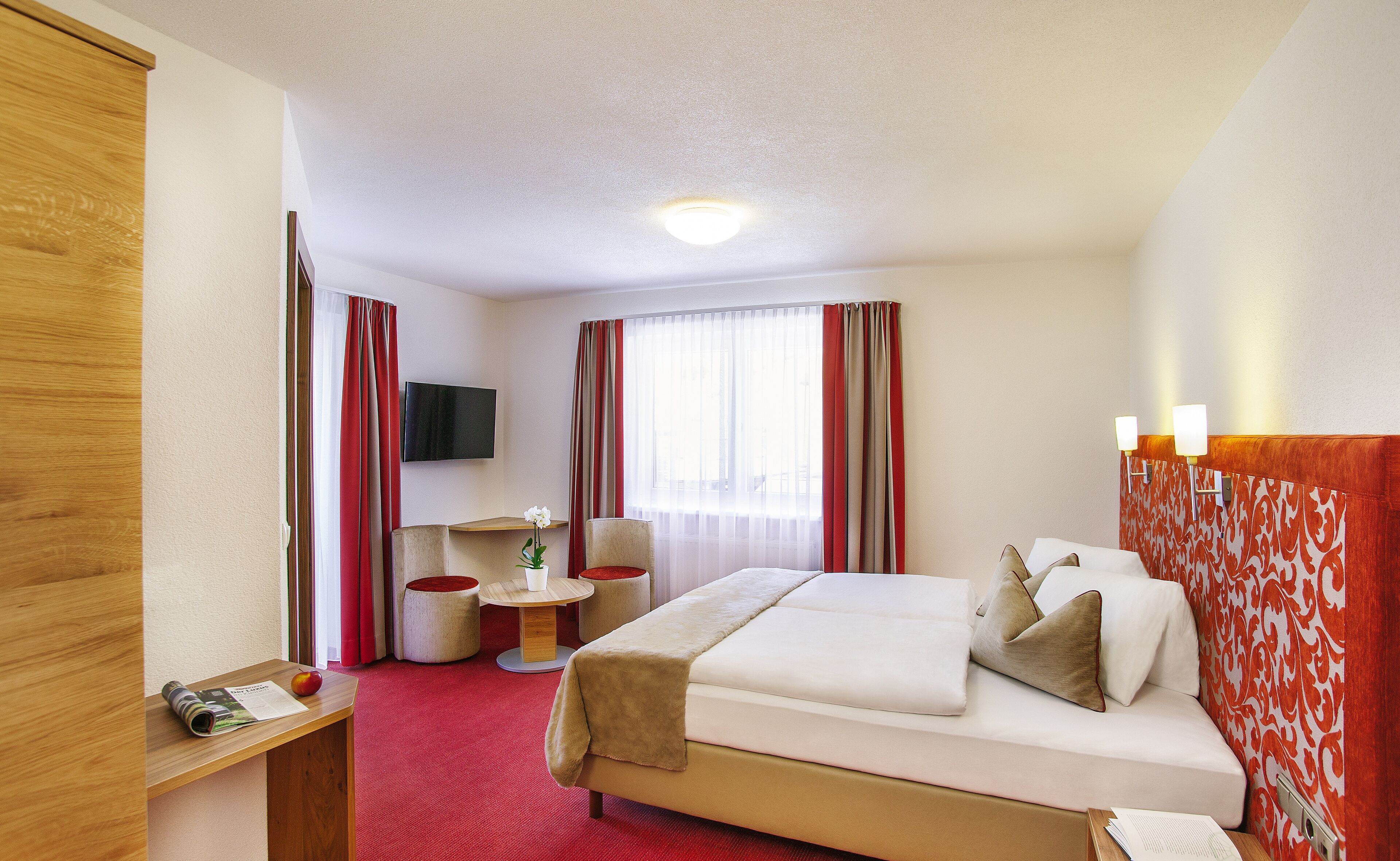 A hotel room with a bed, a desk, a chair, a small table, a TV, and a window with curtains.