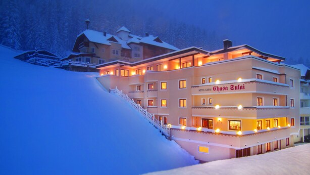 A multi-story hotel with a snow-covered exterior, a staircase leading to the entrance, and illuminated windows. - Hotel Chasa Sulai (Ischgl)