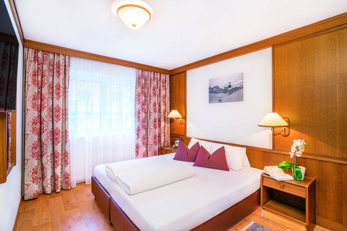 Habitación de hotel con una cama grande, dos lámparas de noche, una mesita de noche con un libro y un jarrón de flores, y una ventana con cortinas estampadas.