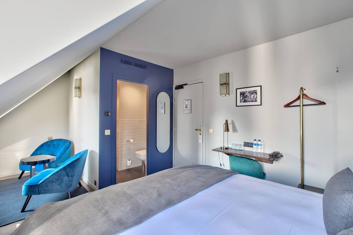 A modern hotel room with a bed, a desk with a chair, a hanging coat rack, and a bathroom visible through an open door.