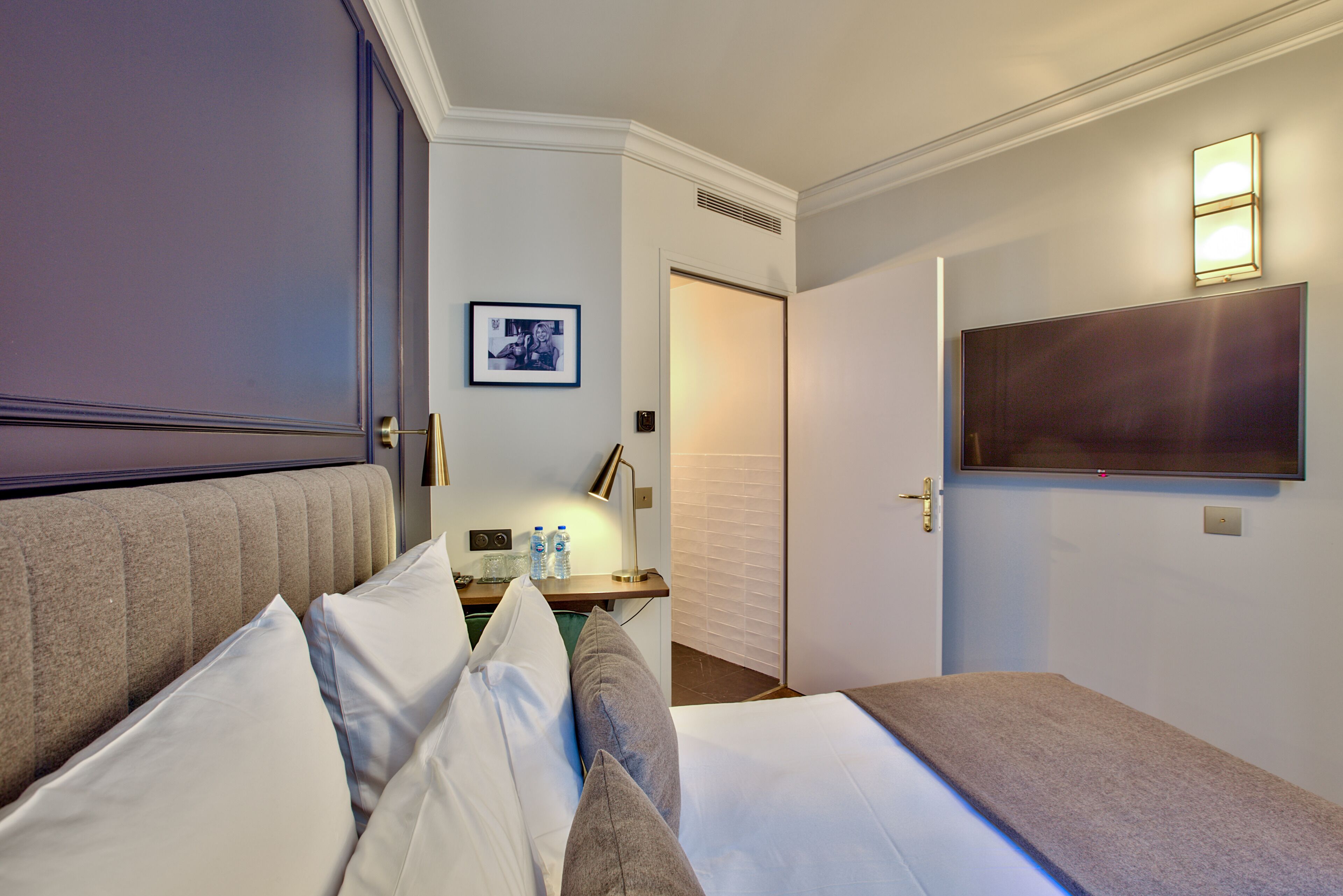 A hotel room with a large bed, a bedside table with a lamp, a flat-screen TV mounted on the wall, and a framed picture on the wall.