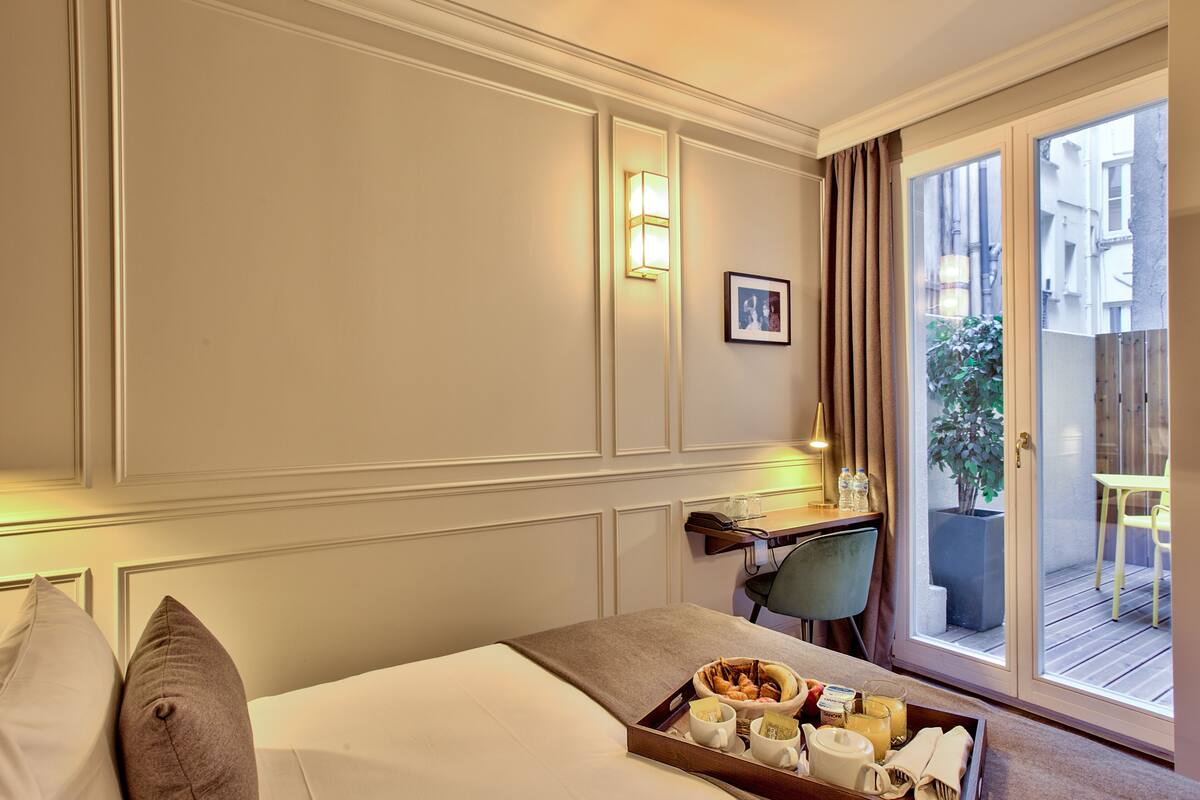 A hotel room with a bed, a tray of breakfast items, a desk with a chair, a window with curtains, and a potted plant outside.
