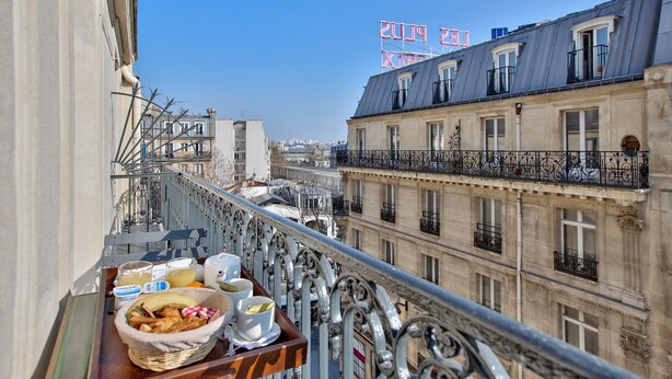Un balcon avec un plateau de mets pour le petit-déjeuner, donnant sur un paysage urbain avec des bâtiments historiques et un ciel bleu dégagé.