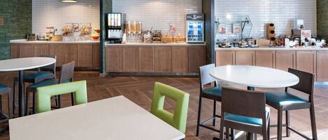 A modern cafe with wooden cabinets, a central counter with various food items, and round tables with chairs.