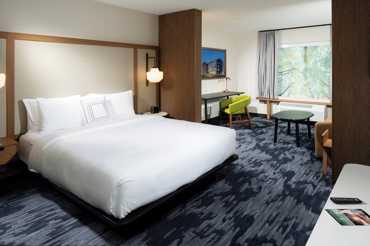 Ein Hotelzimmer mit einem großen Bett, einem Schreibtisch mit Stuhl, einem kleinen Tisch und einem Fenster mit Blick auf Gebäude.