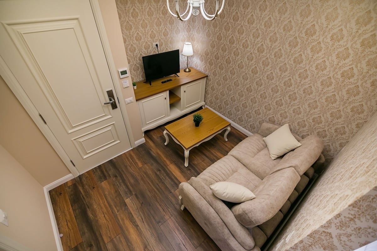A living room with a beige sofa, a wooden coffee table, a TV on a wooden stand, and patterned wallpaper.