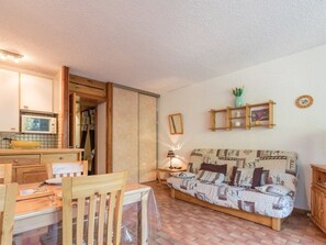 Ein gemütliches Wohnzimmer mit einem Holzesstisch, einer Couch mit gemusterten Kissen, einem kleinen Holzregal mit Dekoration und einer Mikrowelle auf der Arbeitsfläche.