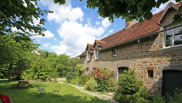 A stone house with a garden, a picnic table, and two red lounge chairs.