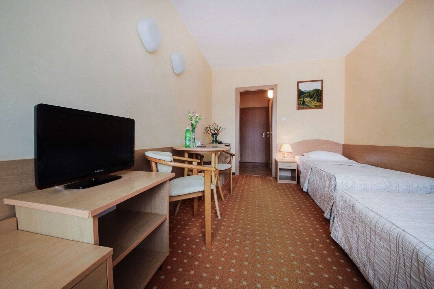 Habitación de hotel con cama, escritorio con televisor, silla, una mesita con un florero y puerta que da a otra habitación.
