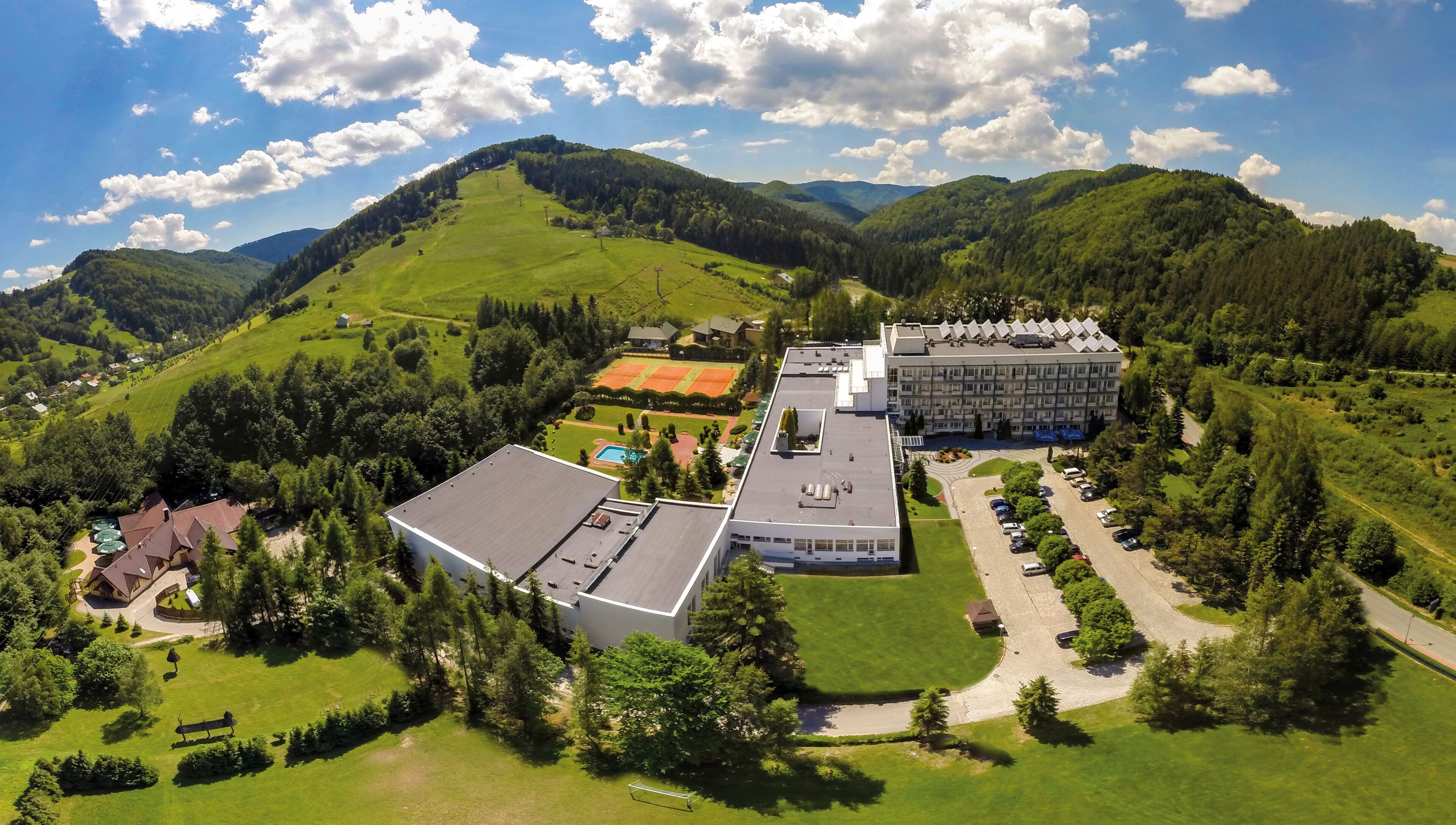 An aerial view of a hotel with a swimming pool, tennis courts, and a parking area surrounded by lush green hills and trees.