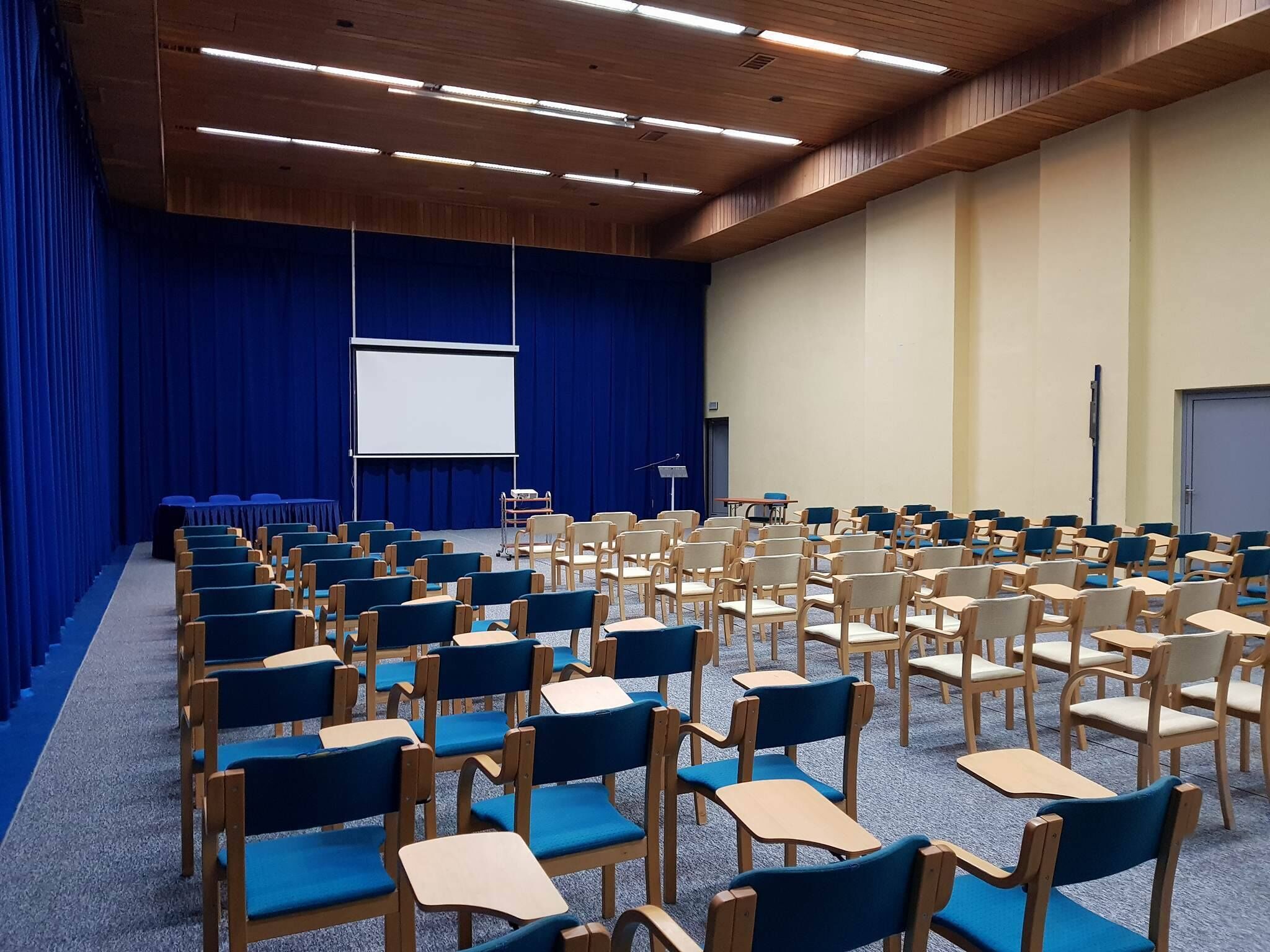 Un salón de conferencias con filas de sillas y mesas, una pantalla de proyector y una zona de escenario.