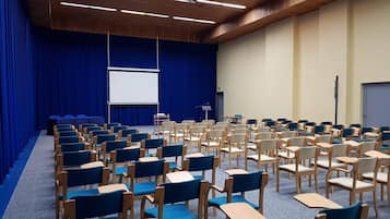 Un salón de conferencias con filas de sillas y mesas, una pantalla de proyector y una zona de escenario.
