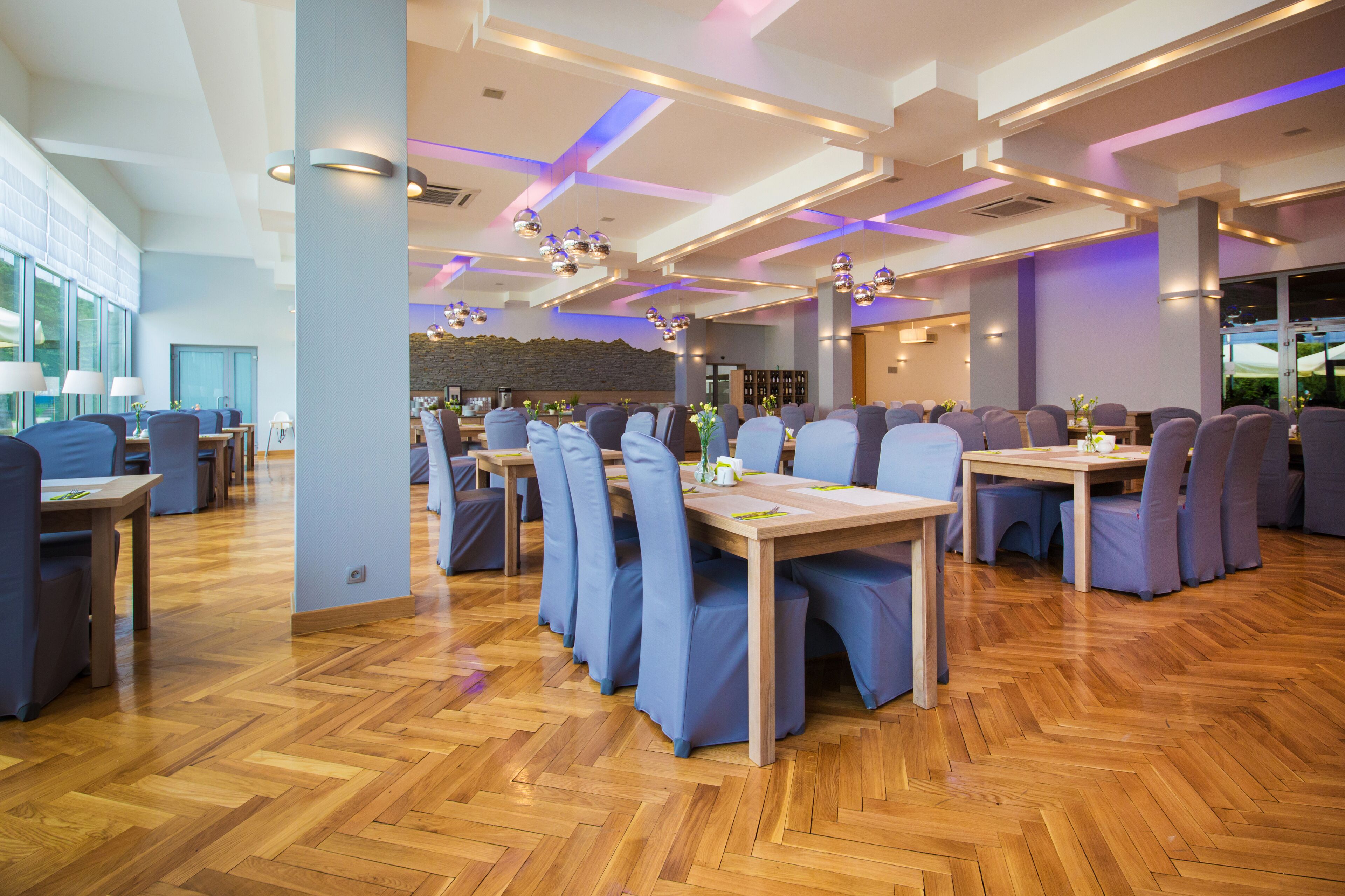 A spacious restaurant with wooden flooring, blue upholstered chairs, and pendant lighting.