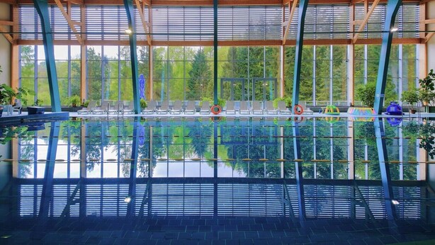 Una piscina interior con paredes de vidrio transparente, vigas de madera y vistas a árboles fuera.