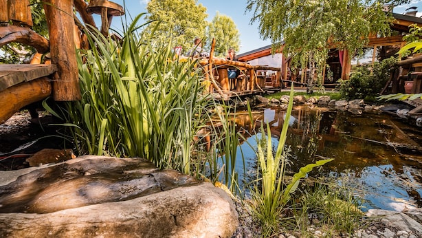 Eine rustikale Holzbrücke über einen Teich, im Hintergrund ein Holzenstall.