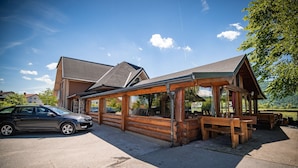 Eine Holzhütte mit überdachter Terrasse, ein geparktes Auto davor und eine Bergkette im Hintergrund.