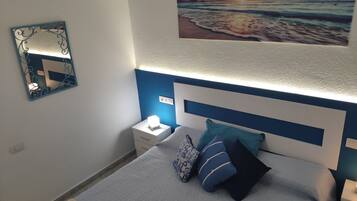 Een slaapkamer met een bed, nachtkastje, spiegel aan de muur en een schilderij van een strand.