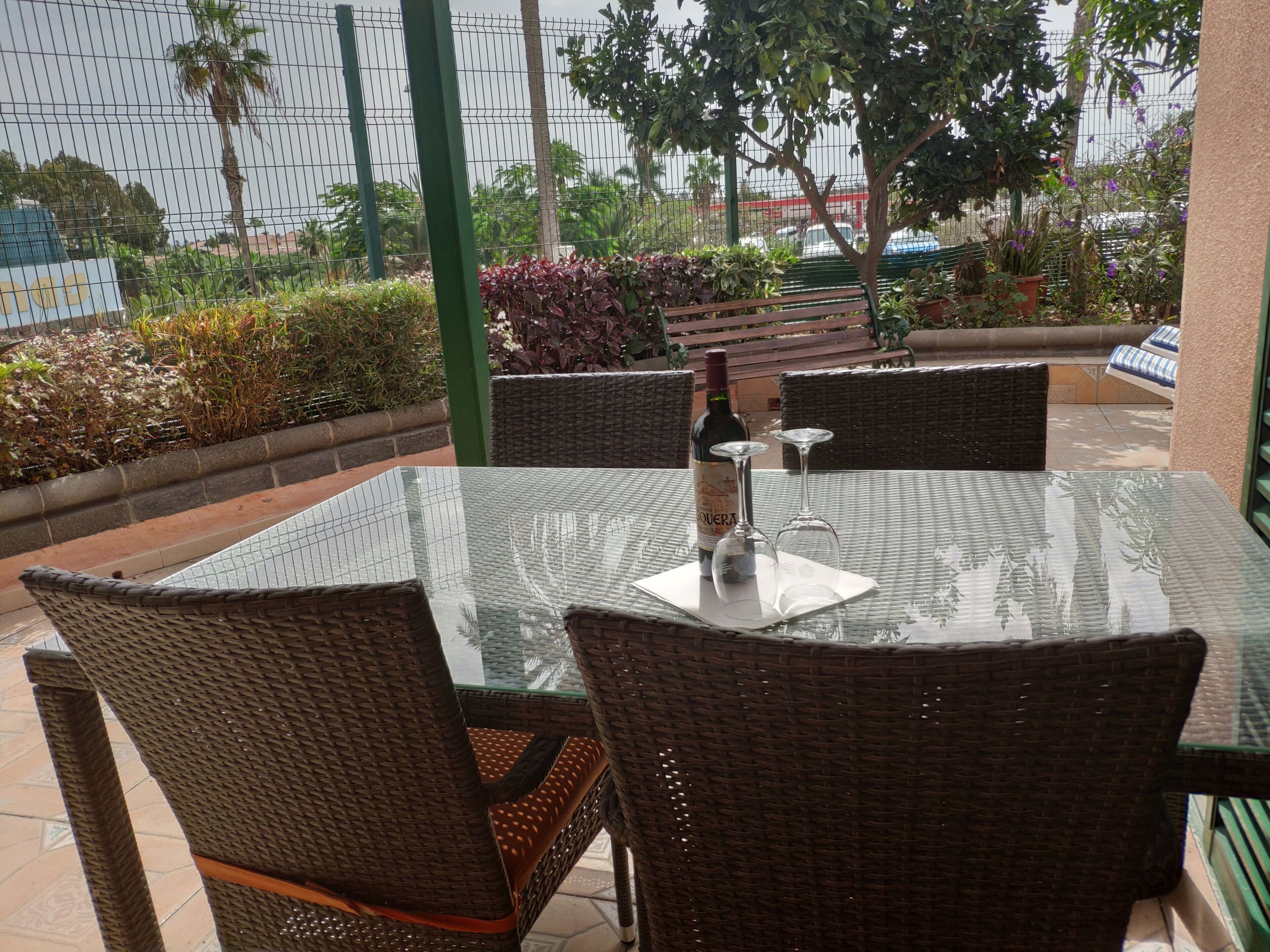 Een terras buiten met rieten stoelen, een glazen tafel, een fles wijn en een glas.