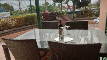 Een terras buiten met rieten stoelen, een glazen tafel, een fles wijn en een glas.
