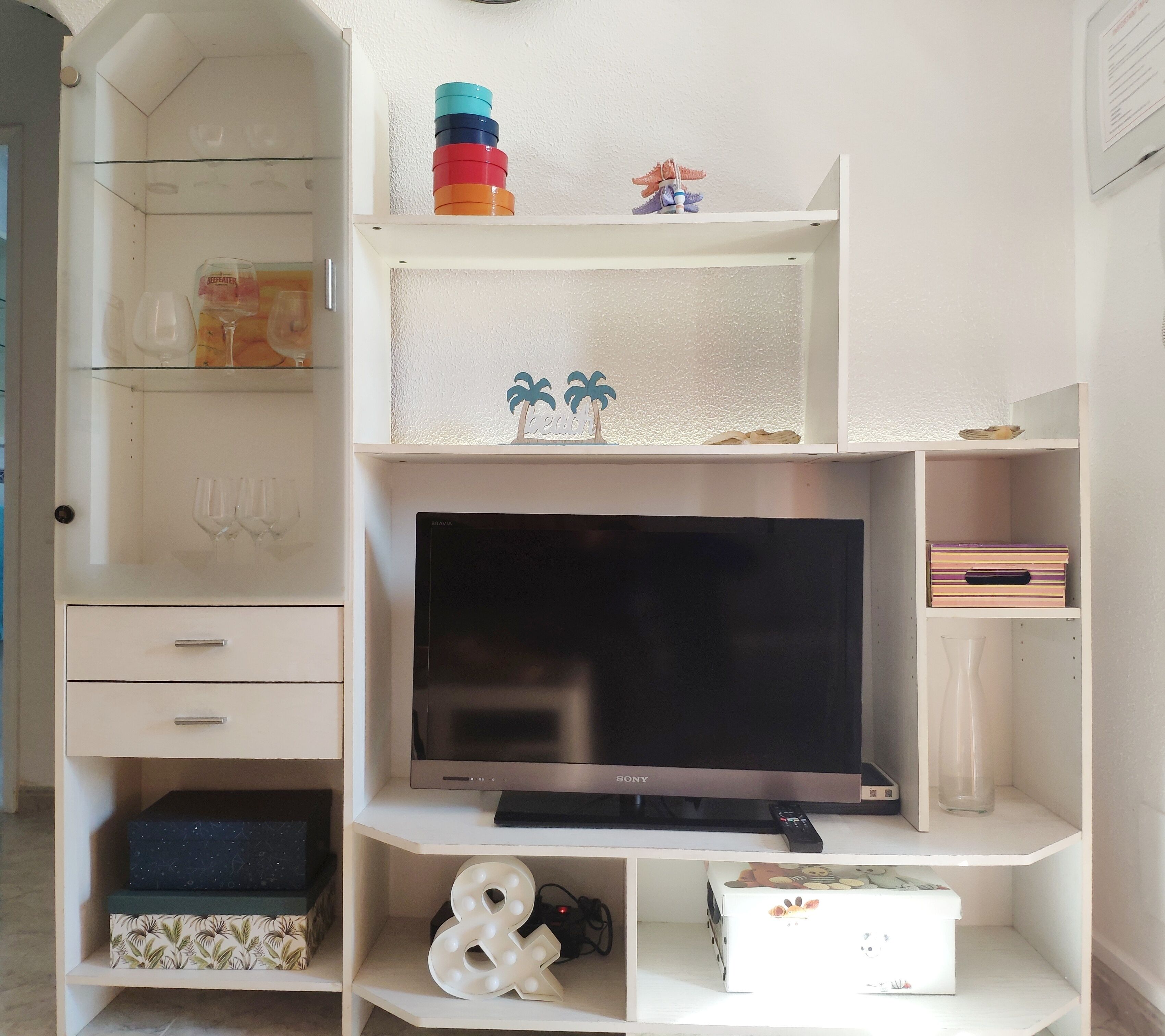 Un meuble de rangement blanc pour appareils multimédias, avec un téléviseur à écran plat, des étagères ornées d’objets décoratifs et une petite table sur laquelle repose une télécommande.