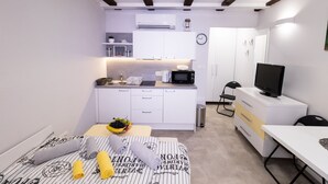 Een compacte hotelkamer met een kitchenette, een bed met gestreept beddengoed, een tafeltje met een schaal fruit en een televisie op een standaard.