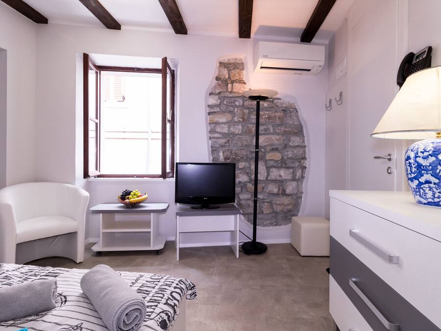 A bedroom with a bed, a TV, a small table with a fruit bowl, a white chair, a lamp, and a dresser with a vase.