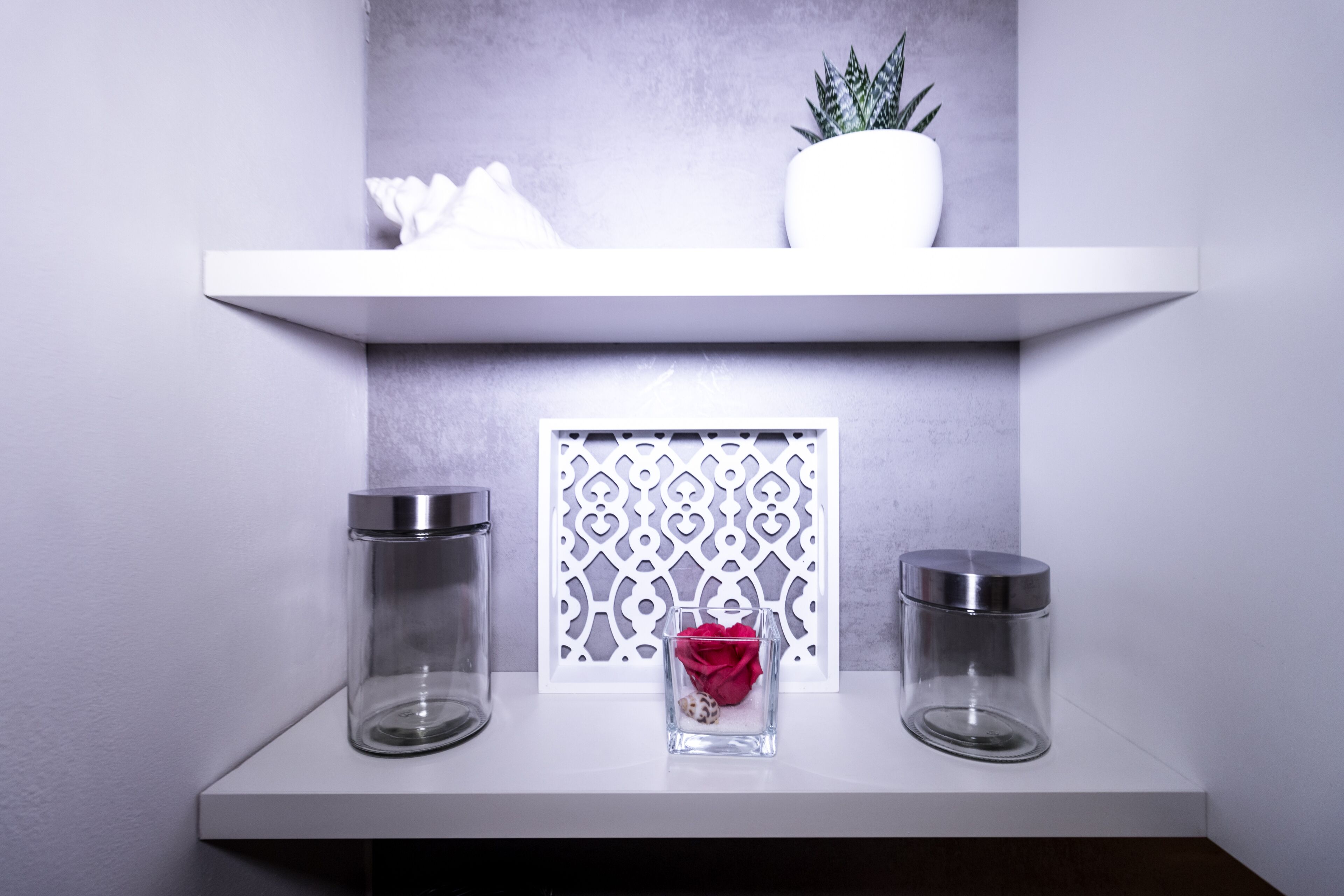 A white shelf with two glass jars and a small vase with a red flower.