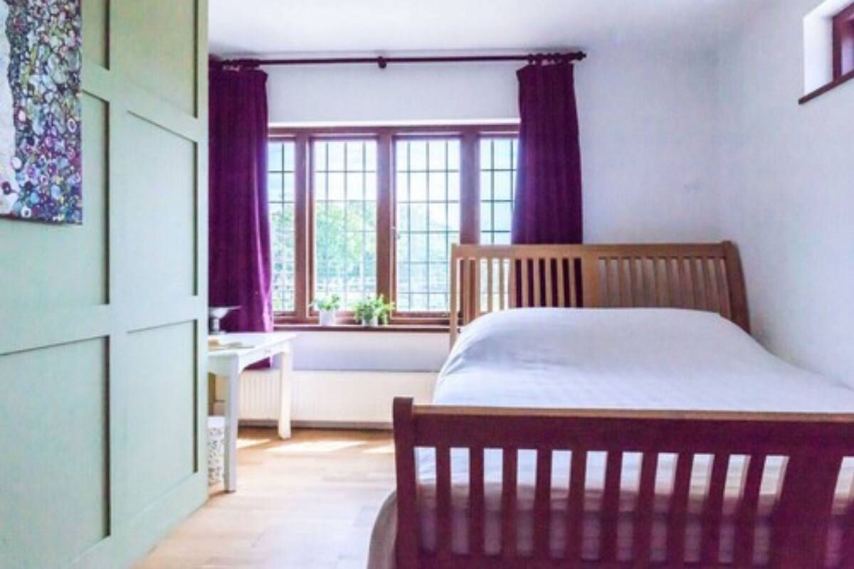 A bedroom with a wooden bed, a window with curtains, a radiator, and a small table with a plant.