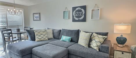 Ein Wohnzimmer mit grauem Ecksofa, einem Esstisch mit Stühlen, einem Deckenventilator und Wandbildern.