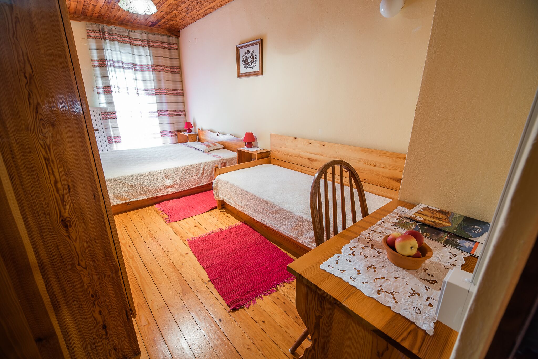 A cozy bedroom with two beds, a wooden table with a bowl of fruit, and a red rug on the floor.